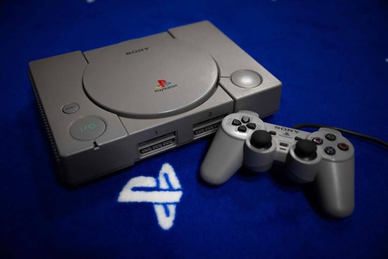 A imagem mostra um console PlayStation original da Sony, de cor cinza, posicionado sobre um fundo azul. O console possui um botão de liga/desliga e duas portas para controle, além do logotipo do PlayStation visível na parte superior. Ao lado do console, há um controle cinza com botões coloridos e analógicos.