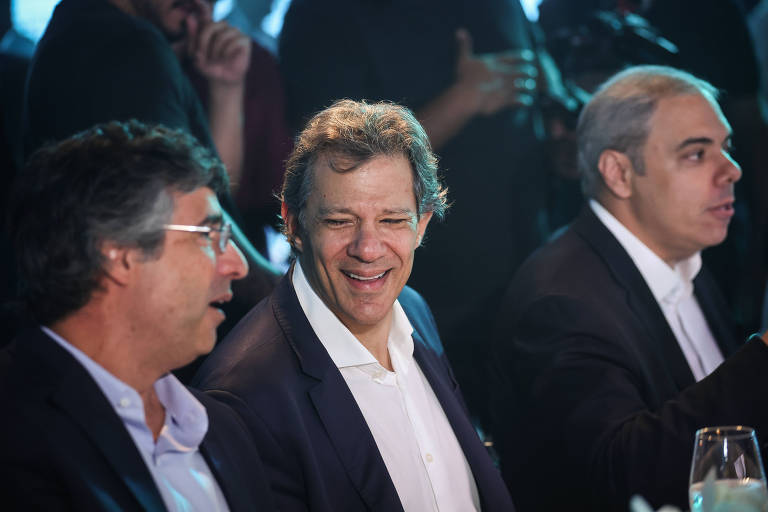 A imagem mostra três homens sentados em uma mesa durante um evento social. O homem ao centro, com cabelo castanho e um sorriso, parece estar conversando com os outros dois. À esquerda, um homem com cabelo grisalho e óculos, e à direita, um homem com cabelo curto e grisalho. O ambiente é iluminado com luzes azuis ao fundo.