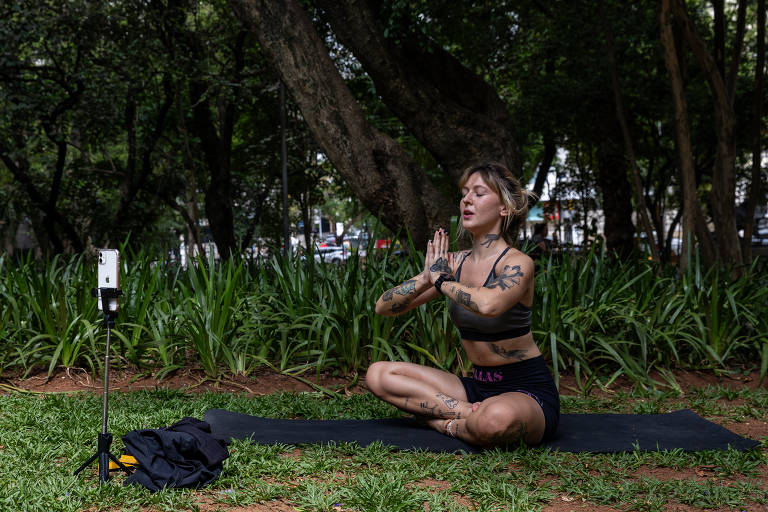 Uma mulher branca, de cabelos claros, jovem e com taguagens nos braços, barriga, pescoço e pernas, está sentada ao centro da imagem; ela está sentada, com pernas cruzadas, sobre uma toalha escura na grama, e junta as mãos abertas à frente do rosto; o entorno é repleto de plantas e árvores