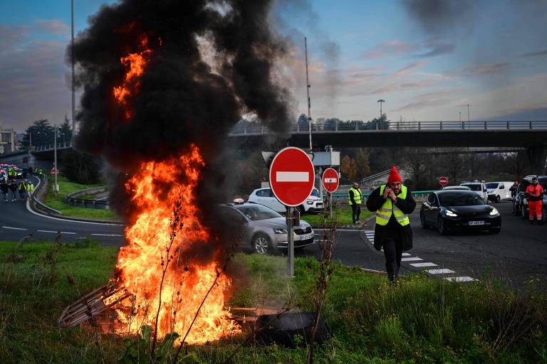 A imagem mostra um incêndio em uma barricada na beira de uma estrada. Chamas altas e fumaça preta se elevam, enquanto um sinal de trânsito de proibição de entrada está visível ao lado. Algumas pessoas vestindo coletes amarelos estão presentes, e veículos estão parados na estrada ao fundo.