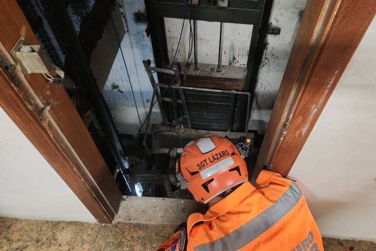 Um técnico vestido com um uniforme laranja e um capacete está agachado em frente a um poço de elevador. O interior do poço é visível， mostrando as estruturas metálicas e cabos do elevador. As paredes ao redor são de cor clara e o chão é de madeira.