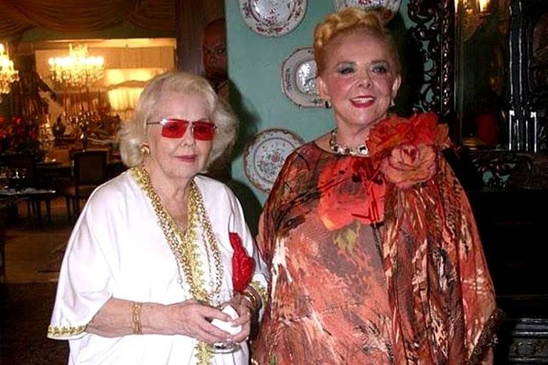 Em foto colorida, duas mulheres de vestido branco e vestido estampado posam para foto