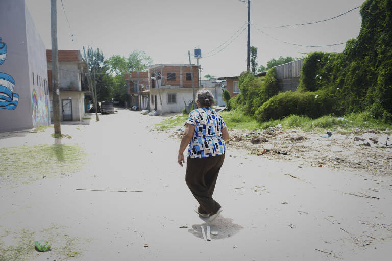 A imagem mostra uma mulher idosa de costas, caminhando em uma rua de um bairro. Ela usa uma blusa colorida com padrões e calças escuras. Ao fundo, há casas de dois andares e um muro com grafite. O chão está coberto de detritos e a vegetação é visível nas laterais da rua. O céu está claro e a iluminação é natural.