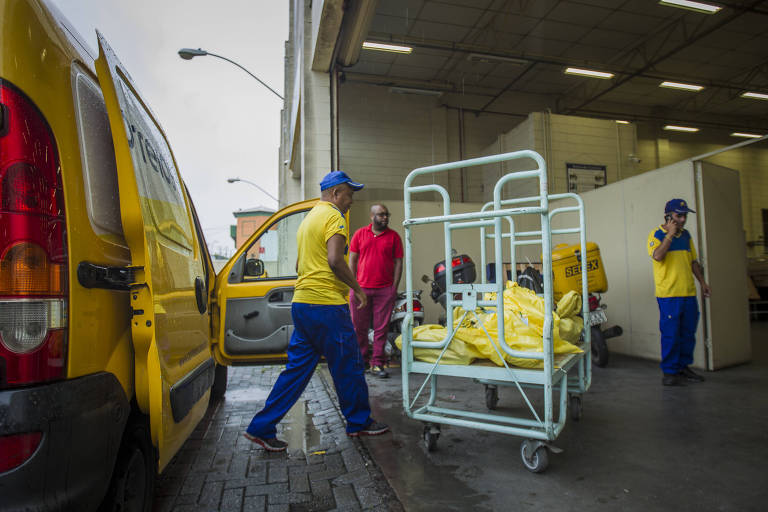 A imagem mostra um ambiente de trabalho onde três pessoas estão envolvidas em atividades de carga e descarga. Um homem está puxando um carrinho de metal com sacos amarelos， enquanto outro homem， vestido de vermelho， observa. Um terceiro trabalhador está dentro de uma van amarela， que está parcialmente aberta. O local parece ser uma área de entrega， com um piso de concreto e paredes claras.