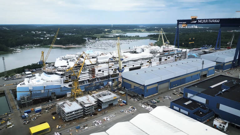 Imagem aérea de um estaleiro com vários navios em construção. Há guindastes amarelos e estruturas metálicas visíveis. Ao fundo, pode-se ver um corpo d'água e uma área verde. O céu está parcialmente nublado.