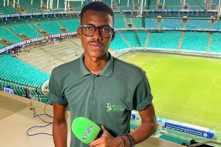 Um jovem apresentador está posando em um estúdio de transmissão, segurando um microfone verde. Ele usa uma camisa polo verde e está em frente a um campo de futebol iluminado, com arquibancadas vazias ao fundo.