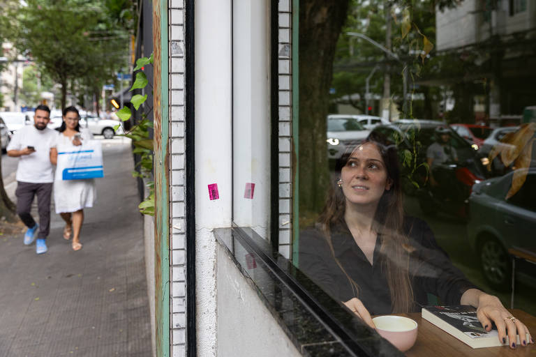 A imagem mostra uma mulher sentada em uma mesa, olhando para fora através de uma janela. Ela segura uma xícara e parece pensativa. Do lado de fora, duas pessoas caminham na calçada, uma delas carregando uma caixa. O ambiente é urbano, com árvores e carros visíveis ao fundo.