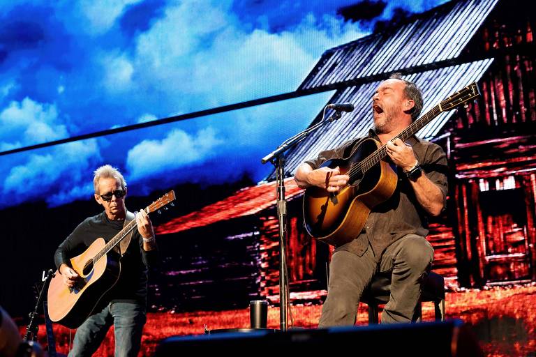 Dois músicos se apresentam em um palco, um deles toca violão e o outro canta enquanto toca guitarra. Ao fundo, há uma projeção de uma casa de campo com um céu azul e nuvens.