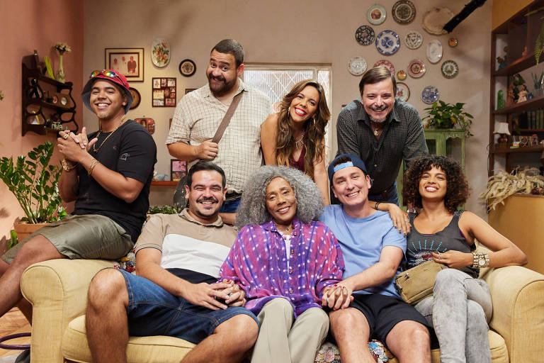 A imagem mostra um grupo de oito pessoas sorrindo em um ambiente interno, possivelmente uma sala de estar. Eles estão sentados e em pé ao redor de um sofá amarelo. A mulher no centro, com cabelo grisalho e uma blusa colorida, segura as mãos de dois homens ao seu lado. Os outros membros do grupo estão distribuídos ao fundo e à frente, todos com expressões alegres. O ambiente é decorado com plantas e objetos pessoais nas paredes.