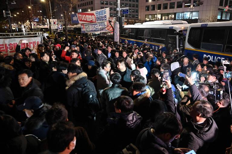 Multidão com celulares ao lado de ônibus da polícia