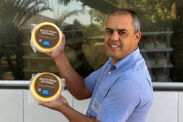 Um homem está posando para a foto， segurando dois recipientes redondos com tampas. Ele está sorrindo e vestindo uma camisa azul. Ao fundo， há uma janela e árvores visíveis， sugerindo um ambiente externo.