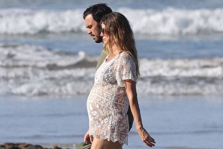 Um casal caminha na praia, com ondas ao fundo. A mulher está grávida, vestindo uma blusa de renda branca e shorts. O homem usa uma camiseta verde e shorts. Ambos estão descalços, e a areia está molhada, refletindo suas sombras.