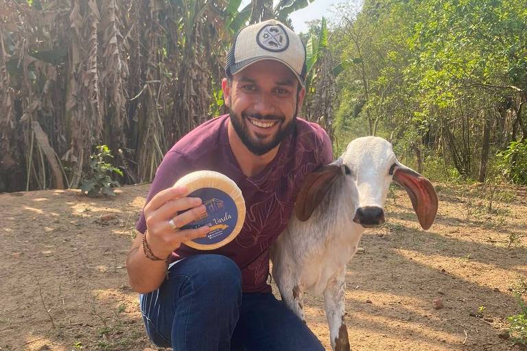 Um homem está agachado em um terreno rural， segurando um objeto redondo na mão. Ao seu lado， há uma cabra com orelhas longas. O fundo apresenta vegetação， incluindo árvores e plantas， sugerindo um ambiente natural e rural.