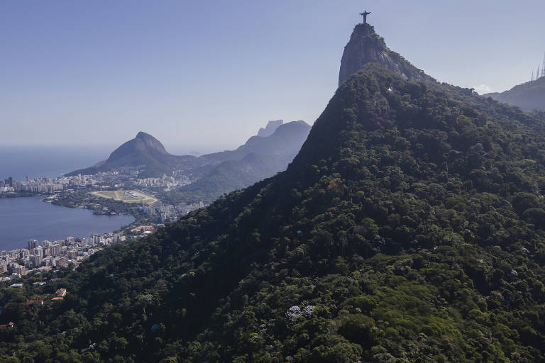 A imagem mostra uma vista panorâmica do Rio de Janeiro， destacando o Cristo Redentor no topo do Corcovado， cercado por montanhas verdes. Ao fundo， é possível ver o Pão de Açúcar e a baía da Guanabara， com a cidade se estendendo ao longo da costa sob um céu claro.
