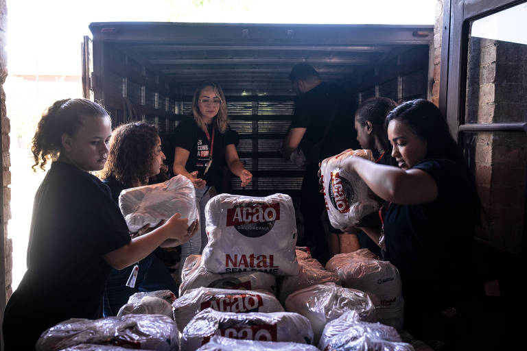 A imagem mostra um grupo de pessoas trabalhando em um caminhão， descarregando pacotes de alimentos. Algumas pessoas estão dentro do caminhão， enquanto outras estão do lado de fora， organizando os pacotes. Todos estão usando camisetas pretas e parecem concentrados na tarefa. Os pacotes têm a marca 039;Ação da Cidadania039; e a frase 039;Natal Sem Fome039;.