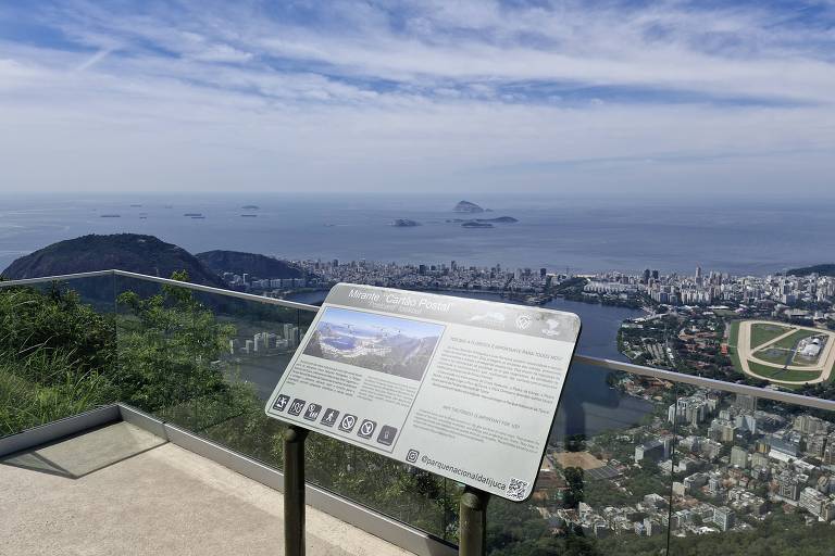 A imagem mostra uma vista panorâmica do Rio de Janeiro， com o mar ao fundo e algumas ilhas visíveis. Em primeiro plano， há um painel informativo em uma estrutura de metal， que parece descrever a vista. O céu está parcialmente nublado， e a cidade se estende ao longo da costa.