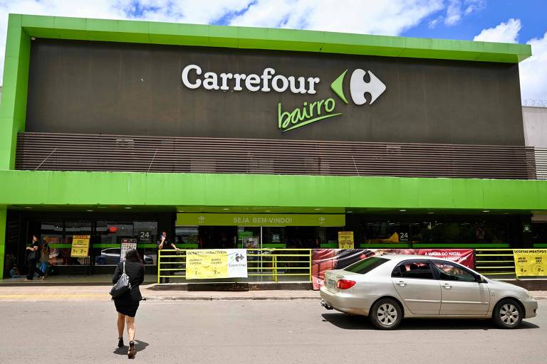 A imagem mostra a fachada de uma loja Carrefour Bairro, com uma grande placa verde e o logotipo do Carrefour. Na frente da loja, há um carro estacionado e uma pessoa caminhando em direção à entrada. O céu está claro e azul.
