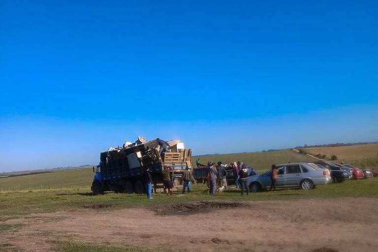 A imagem mostra um caminhão grande carregado com diversos objetos, incluindo paletes e caixas, estacionado em um campo aberto. Há várias pessoas ao redor do caminhão, algumas parecendo ajudar ou observar. Ao fundo, há um céu azul claro e uma área verde, com alguns carros estacionados em uma linha. O terreno é de terra e grama.