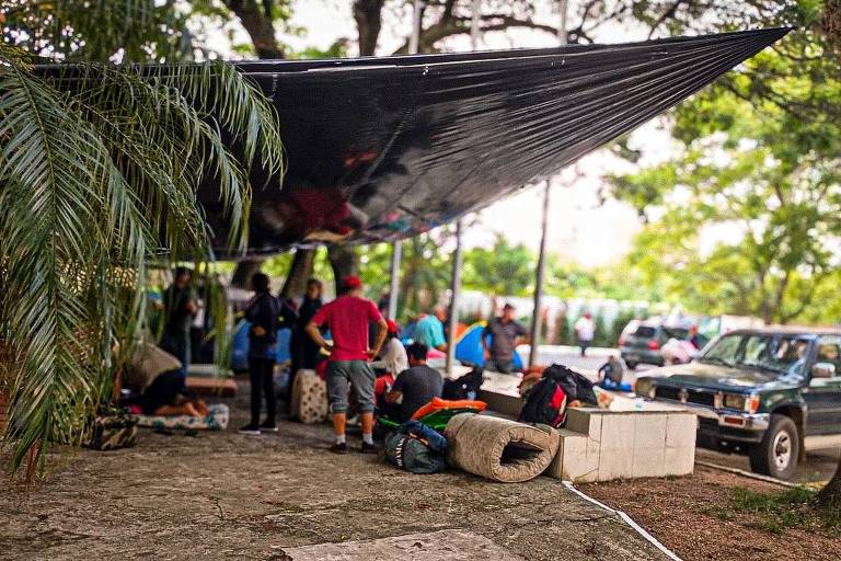 A imagem mostra um grupo de pessoas reunidas sob uma grande cobertura preta, com árvores ao fundo. Algumas pessoas estão agachadas, aparentemente organizando ou manipulando equipamentos, enquanto outras estão em pé. Há mochilas e objetos espalhados pelo chão. Um veículo está estacionado próximo ao local, e a cena é cercada por vegetação.