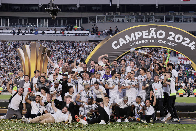 Um grupo de jogadores e membros da equipe celebra a conquista da Copa Libertadores 2024. Eles estão reunidos em frente a um troféu dourado, com um grande banner acima que diz 'CAMPEÃO 2024 CONMEBOL LIBERTADORES'. A cena é festiva, com muitos sorrisos e gestos de alegria, enquanto alguns seguram copos e outros fazem poses para fotos. O fundo é preenchido por torcedores nas arquibancadas, vestindo camisas listradas.
