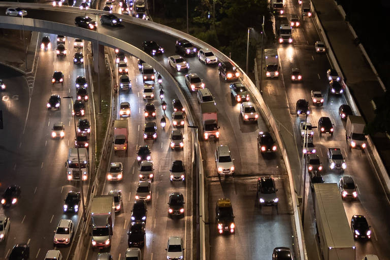 Imagem mostrando uma vista aérea de um grande fluxo de tráfego em uma rodovia à noite， com muitos carros e caminhões iluminados por faróis. A estrada tem curvas e várias faixas， evidenciando a densidade do tráfego.