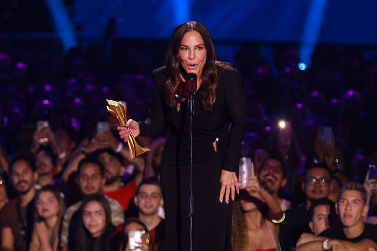 Uma mulher em um vestido preto está em um palco, segurando um prêmio dourado e falando em um microfone. Ao fundo, uma multidão de pessoas aplaude e grava com seus celulares. O ambiente é iluminado com luzes azuis e roxas.