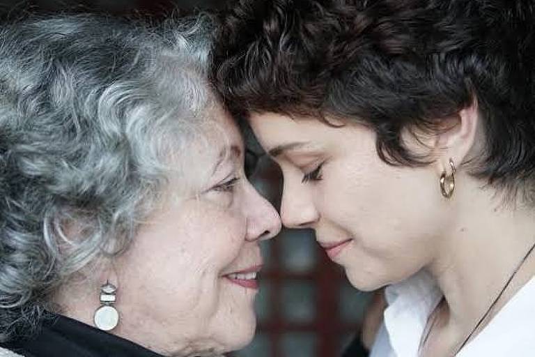 A imagem mostra duas mulheres de rostos próximos, compartilhando um momento íntimo. A mulher mais velha tem cabelo grisalho e usa um suéter claro, enquanto a mulher mais jovem tem cabelo curto e castanho, vestindo uma camisa branca. Ambas estão sorrindo suavemente, com expressões de afeto e carinho. O fundo é desfocado, destacando a conexão entre elas.