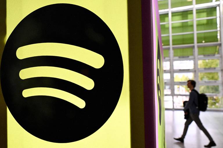 A imagem mostra o logotipo do Spotify， que consiste em três linhas curvas em um círculo preto， em um fundo amarelo. Ao lado， uma pessoa com uma mochila caminha em direção a uma entrada， com uma estrutura moderna e janelas ao fundo.