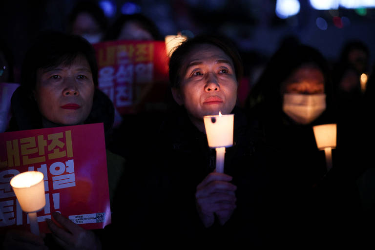 A imagem mostra um grupo de pessoas em uma manifestação à noite, segurando velas acesas e cartazes. As pessoas estão em um ambiente escuro, com algumas expressões sérias. Uma mulher em destaque está segurando uma vela e olhando para frente, enquanto outras ao fundo seguram cartazes com texto em coreano.