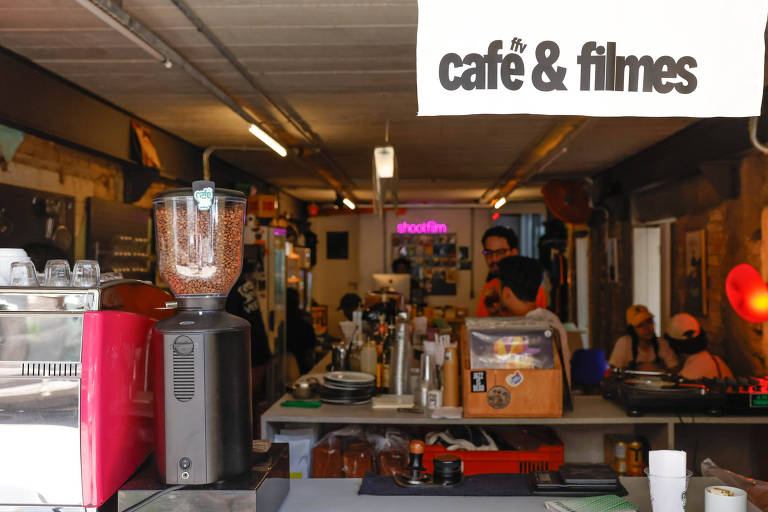 A imagem mostra o interior de um café com uma área de atendimento. No fundo， há um balcão com uma máquina de café， um liquidificador e outros equipamentos. Um letreiro suspenso diz 039;café amp; filmes039;. Algumas pessoas estão visíveis no local， e a iluminação é suave， com algumas luzes coloridas ao fundo.