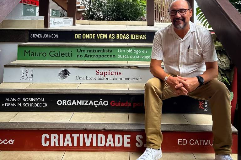 Um homem está sentado em uma escada que é decorada com a aparência de livros. Ele usa uma camisa branca e calças bege， com sapatos brancos. As escadas têm várias inscrições em cores diferentes， incluindo 039;Criatividade039;， 039;Organização039;， e nomes de autores como 039;Mauro Galetti039;. Ao fundo， há vegetação e uma estrutura de madeira.