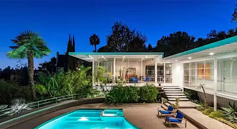 A imagem mostra uma casa moderna com uma piscina em primeiro plano. A casa possui grandes janelas e uma varanda, cercada por vegetação tropical. À esquerda, há uma palmeira e outras plantas. O céu está claro e azul, indicando que é ao entardecer.