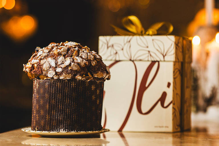 A foto mostra um panetone com massa coberta com chocolate e praliné de castanha de caju. Ao fundo， uma bonita caixa onde se lê Oli