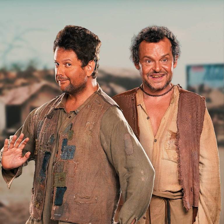 A imagem mostra dois homens em um cenário rural. O homem à esquerda está sorrindo e acenando, vestindo uma camisa e um colete com remendos coloridos. O homem à direita também está sorrindo, usando uma camisa aberta e um colete de tecido rasgado. Ao fundo, há uma paisagem com casas simples e um céu claro