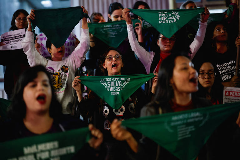 A imagem mostra um grupo de pessoas em uma manifestação， segurando lenços verdes. Algumas pessoas estão gritando e expressando suas emoções. Os lenços têm inscrições que incluem 039;JUSTIÇA039; e 039;SALVA VIDAS039;. O ambiente parece ser um espaço público， com outras pessoas ao fundo também participando do ato.