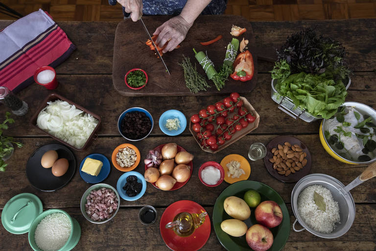 A imagem mostra uma mesa de cozinha com diversos ingredientes dispostos. Uma mão está cortando cenouras em uma tábua de corte. Há uma variedade de vegetais， incluindo tomates， cebolas， alface e ervas. Também estão presentes ovos， grãos， frutas como maçãs e uma tigela com arroz. Os ingredientes estão organizados em recipientes coloridos e há um pano de prato ao fundo.