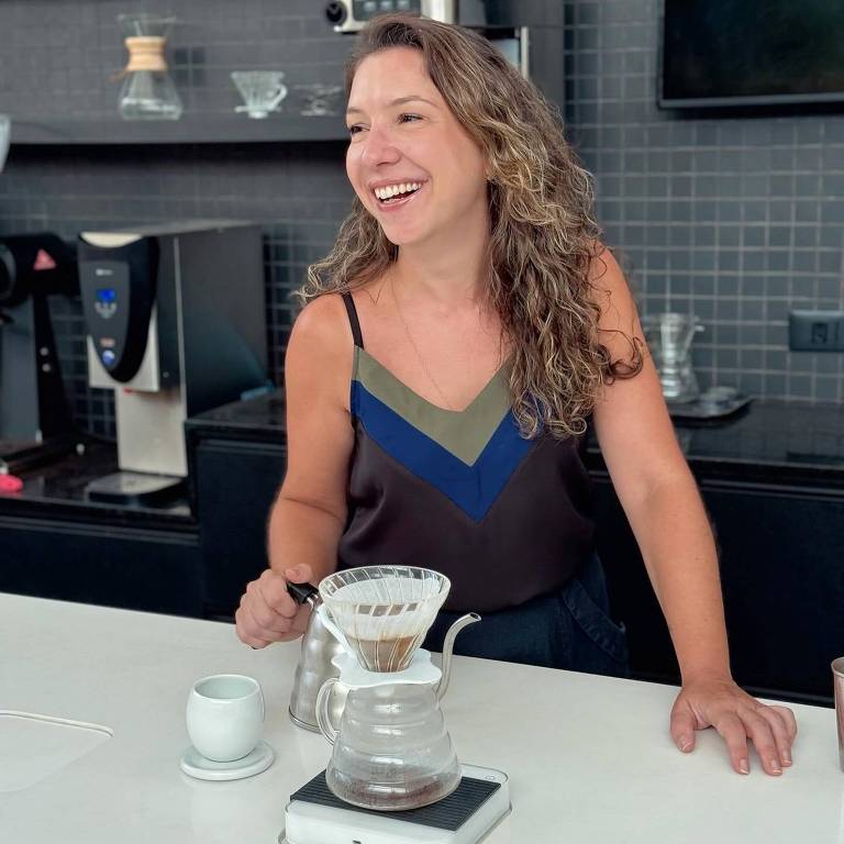 Uma mulher com cabelo cacheado e solto， vestindo uma blusa de alças com cores verde e azul， está sorrindo enquanto prepara café em uma bancada. Ela segura uma jarra de café e está usando um coador de café sobre uma jarra de vidro. Ao lado， há uma xícara branca e um suporte para a jarra. O fundo é uma cozinha moderna com prateleiras e equipamentos de café.