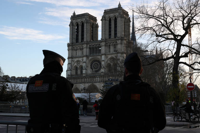 A imagem mostra dois policiais de costas, observando a catedral de Notre-Dame em Paris. A catedral, com suas torres góticas e detalhes arquitetônicos, está ao fundo sob um céu claro. Árvores sem folhas estão visíveis nas laterais, e há uma placa de sinalização ao lado direito.