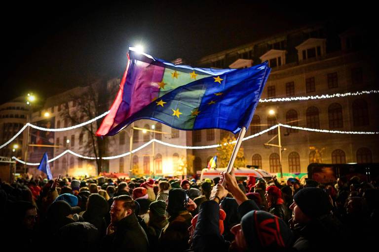 Imagem noturna de uma praça repleta de pessoas e com luzes penduradas. No centro， vê-se as bandeiras da Romênia e da União Europeia segurdas juntas