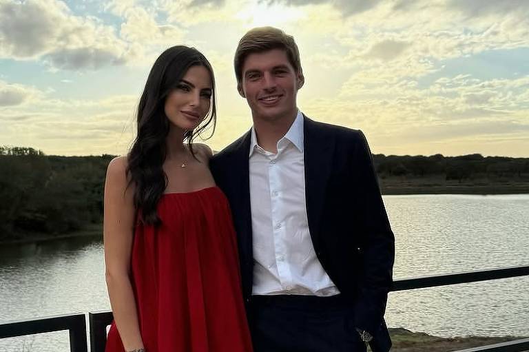 Um casal posando para a foto em um local ao ar livre, com um lago ao fundo. A mulher está vestindo um vestido longo vermelho e segurando uma pequena bolsa vermelha. O homem está usando um terno escuro com uma camisa branca. O céu está parcialmente nublado, com nuvens e um tom dourado ao entardecer.