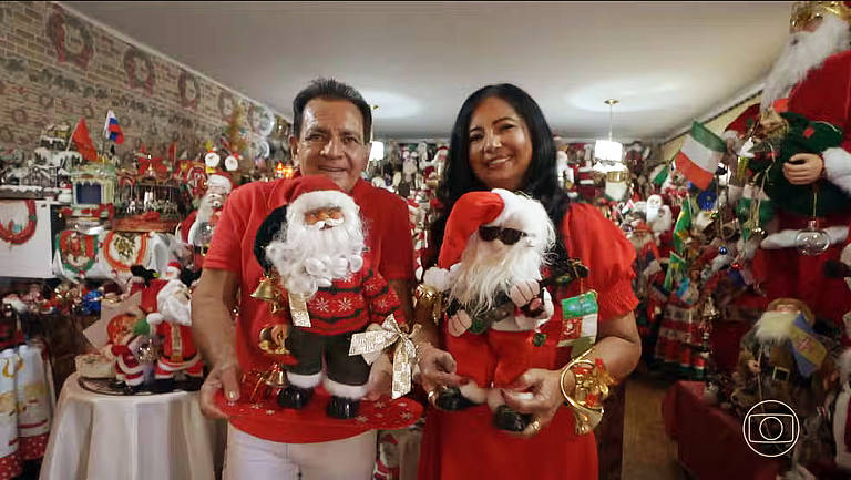 Um casal sorridente está posando em um ambiente decorado para o Natal. Eles seguram dois bonecos de Papai Noel, um com um traje vermelho e outro usando óculos escuros. O fundo é repleto de enfeites natalinos, incluindo outros bonecos de Papai Noel e decorações coloridas. Ambos estão vestidos com roupas vermelhas, e o ambiente é festivo e alegre.