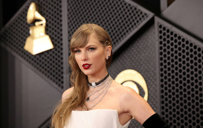 Imagem de uma mulher com cabelo longo e liso, usando um vestido branco e acessórios de prata. Ela está em um evento de premiação, com um fundo que apresenta um troféu de gramofone. A mulher tem lábios vermelhos e uma expressão confiante.