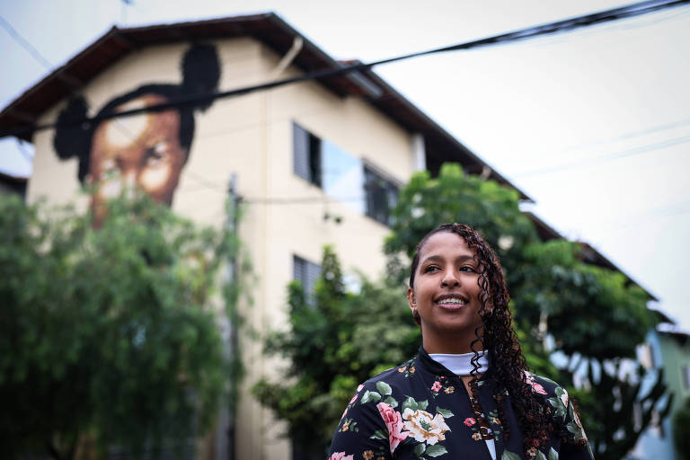 Uma mulher com cabelo cacheado e solto está em primeiro plano， sorrindo. Ela usa uma blusa preta com estampas florais. Ao fundo， há um edifício com um grande mural que retrata um rosto feminino， com um coque e o olhar voltado para a frente. O céu está nublado e há árvores ao redor.