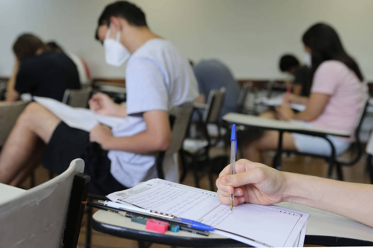 A imagem mostra um grupo de estudantes em uma sala de aula， todos usando máscaras. Eles estão sentados em mesas， concentrados em suas provas. Em primeiro plano， uma mão segura uma caneta， escrevendo em uma folha de papel. Ao fundo， outros alunos também estão escrevendo， com folhas e materiais de estudo sobre as mesas.