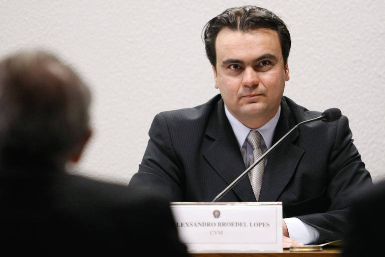 A imagem mostra Alexsandro Broedel Lopes， um homem sentado em uma mesa durante uma audiência pública. Ele está usando um terno escuro e uma gravata clara， com cabelo liso e escuro. À sua frente， há uma placa com o nome 039;ALEXANDRO BRODEL LOPEZ039;. O fundo é de uma parede clara e texturizada. Um outro homem， de costas， está visível à esquerda da imagem.