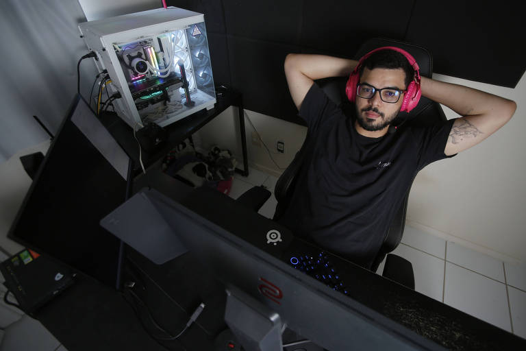 Um homem está sentado em uma cadeira de escritório， com as mãos atrás da cabeça， em um ambiente de trabalho. Ele usa fones de ouvido rosa e óculos. À sua frente， há um monitor de computador e， ao lado， um gabinete de computador com iluminação interna. O fundo é escuro， e o chão é de azulejos brancos.
