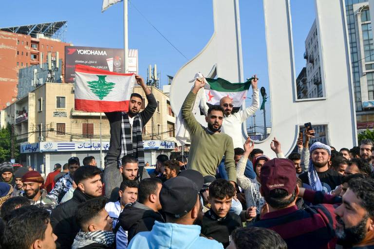 Homens celebram juntos em uma rua. Um deles segura uma bandeira do Líbano, de cabeça para baixo, e outro exibe uma bandeira da Síria.