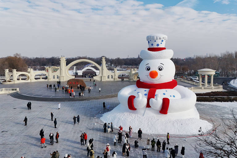 A imagem mostra um grande boneco de neve com um chapéu e um cachecol vermelho, localizado em um parque coberto de neve. Várias pessoas estão ao redor do boneco, que é muito maior do que os visitantes. Ao fundo, há estruturas arquitetônicas e um céu parcialmente nublado.