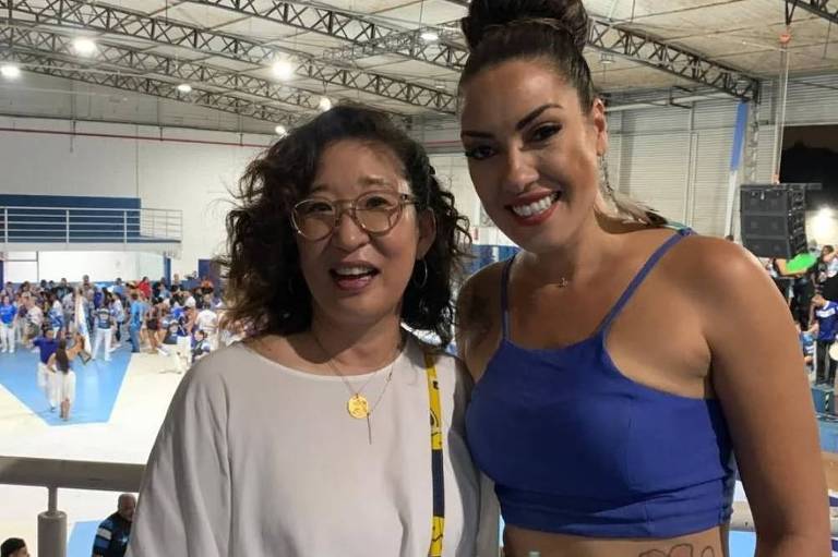 A imagem mostra duas mulheres sorrindo em um evento social. A mulher à esquerda tem cabelo cacheado e usa óculos, vestindo uma blusa branca e uma saia laranja. A mulher à direita tem cabelo preso em um coque e usa uma blusa azul. Ao fundo, há uma multidão de pessoas em um espaço amplo, com piso azul e paredes brancas.