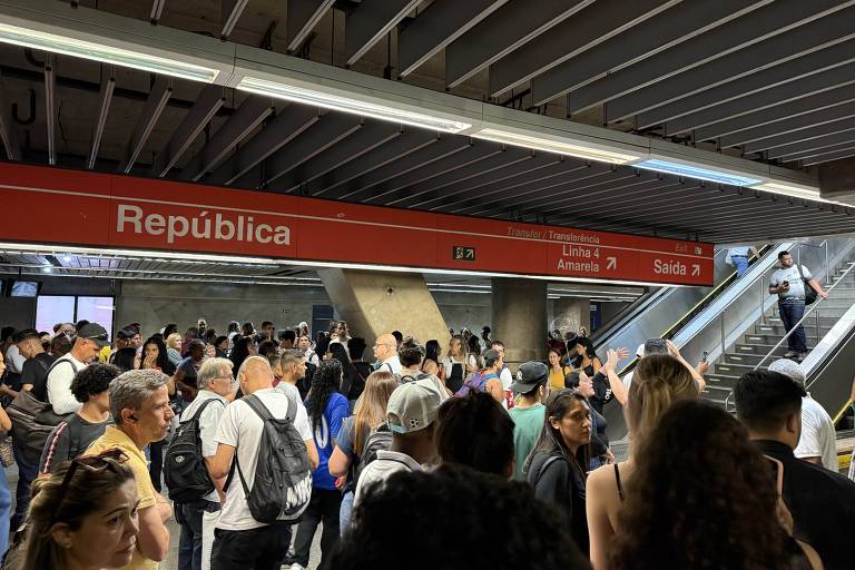 A imagem mostra a estação de metrô República， com um grande número de pessoas na plataforma. O letreiro vermelho indica 039;República039; e há sinalizações para 039;Transferência039;， 039;Linha 4 Amarela039; e 039;Saída039;. Algumas pessoas estão subindo as escadas rolantes， enquanto outras estão paradas， conversando ou aguardando. O ambiente é moderno， com teto de concreto e iluminação artificial.